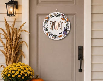 May include: A round white sign with the word SPOOKY in orange text, surrounded by a wreath of autumn leaves, a ghost, a broom, bats, and a purple house. The sign is shown as 12 inches by 12 inches, with other sizes available.
