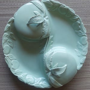 May include: A light green ceramic serving dish with a unique design. The dish is divided into two sections, each featuring a raised peach and leaf motif. The rim is decorated with embossed peaches and leaves. A decorative piece for serving snacks or appetizers.