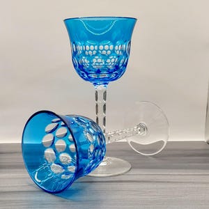 May include: Two blue crystal wine glasses with intricate cut patterns. One glass stands upright, while the other rests on its side. The glasses have a clear stem and base, and the blue bowls feature a detailed design.