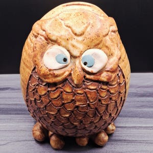 May include: A ceramic owl figurine with a rounded, egg-shaped body. The owl features a textured, brown, scale-like pattern on its chest and a lighter tan, speckled finish on its head. It has two large, protruding white eyes with blue pupils.