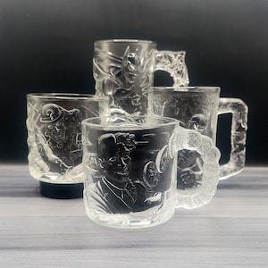 May include: Five clear glass mugs with detailed etched designs. The mugs feature various images, including a person, a figure in a hat, and floral patterns. The handles are also intricately designed, adding to the overall aesthetic.