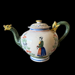 Puede incluir: Una tetera de cerámica blanca con asa y boquilla verdes, adornada con ilustraciones florales y de figuras. La boquilla tiene forma de cabeza de dragón. La tapa y la base presentan detalles en azul y rojo. Una mujer con vestimenta tradicional está pintada en el lateral.