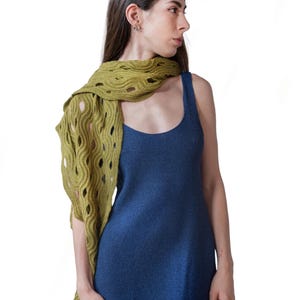 May include: A woman wearing a knitted olive green scarf with a wave-like pattern over a blue dress. The scarf has large, open cutouts. The dress is a solid, dark blue color and has a scoop neckline.