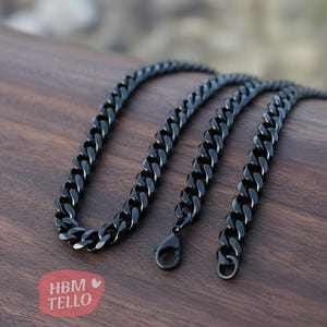 May include: A black curb chain necklace with a lobster clasp. The chain is made of thick, interlocking links and is displayed on a wooden surface. The necklace is a dark, matte black color.