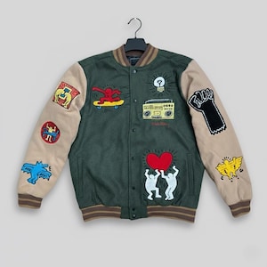 Op de afbeelding: Groene en beige varsity jas met meerdere kleurrijke patches. De jas heeft een rood hart, een skateboarder, een boombox en andere grafische ontwerpen. De mouwen zijn beige en de body is groen met bruine bies.