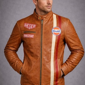 Puede incluir: Chaqueta de cuero marrón con cuello alzado, cierre de cremallera y varios parches. Presenta una franja blanca, roja y naranja en el lado izquierdo y el logotipo "Gulf". La chaqueta tiene dos bolsillos delanteros.