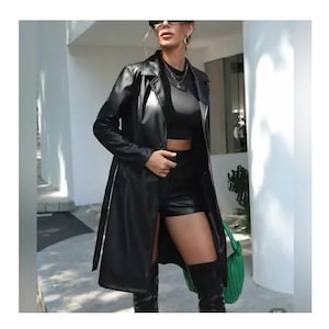 May include: A black faux leather trench coat with a matching belt, worn over a black crop top and shorts. The outfit is completed with knee-high boots and a green handbag. The coat has a classic lapel collar and a sleek, modern design.