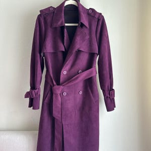 以下が含まれることがあります： ベルト、ボタン、肩章が付いた、プラム色のロングトレンチコート。 袖口にボタンが付いた長袖のコートです。 白い壁を背景に、白いハンガーにコートが掛かっています。
