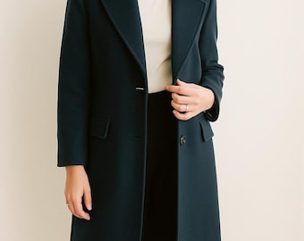 May include: A dark teal overcoat with a classic lapel collar and a single-button closure. The coat has two front pockets and a tailored fit. The model is wearing a white shirt and black pants.