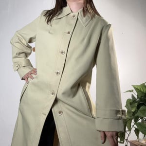 May include: A light green trench coat with a classic design. The coat features a collared neckline, button-down closure, and long sleeves with adjustable cuffs. The coat is made of a smooth fabric and has a tailored fit.