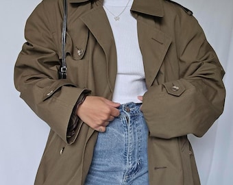 May include: An olive green trench coat with a double-breasted design. The coat features large lapels, button details, and a belt. The model is wearing a white top and blue jeans. A black shoulder bag is visible.