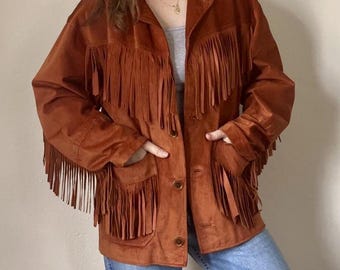 May include: A rust-colored suede jacket with long fringe detailing across the chest, sleeves, and pockets. The jacket has a button-up front and two front pockets. The model is wearing blue jeans and a gray top underneath the jacket.