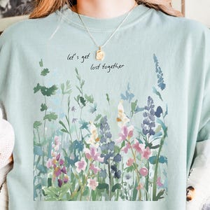 May include: A light blue t-shirt with a watercolor floral design in shades of pink, purple, and green. The text "let's get lost together" is above the floral design. The shirt is part of a clothing collection.