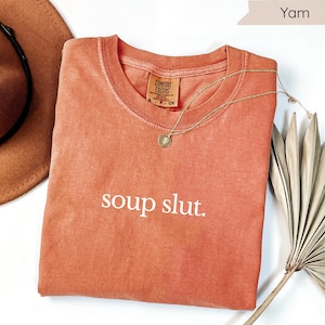 May include: A coral-coloured t-shirt with the phrase "soup slut" printed in white. The shirt has a crew neck and a small tag. A gold necklace and a brown hat are also visible.