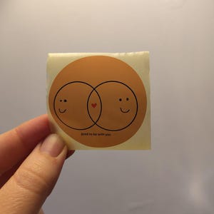 Könnte beinhalten: Ein quadratischer Aufkleber mit hellbraunem Hintergrund. Zwei sich überlappende braune Kreise mit lächelnden Gesichtern und einem roten Herzen in der Mitte. Der Text "good to be with you" steht darunter.
