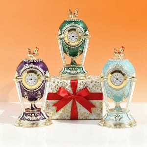 May include: Three decorative egg-shaped clocks in purple, green, and teal. Each clock has a gold-colored frame, a white clock face, and a rooster figurine on top. The clocks are on a gift box with a red bow.