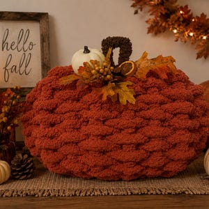 May include: A large, round, rust-colored chunky knit pumpkin decoration featuring a dark brown stem. The top is adorned with a small white pumpkin, dried orange slices, yellow and orange fabric leaves, and small golden berries.