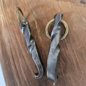 Pode incluir: Dois abridores de garrafa de metal forjados à mão com um design torcido. Cada abridor tem uma extremidade curva e um porta-chaves de latão. Os abridores são exibidos em uma superfície de madeira, mostrando sua estética rústica e artesanato.