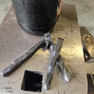 May include: Close-up of hand-forged metal pieces with a twisted design, lying on a metal surface. The pieces are dark gray and have a textured appearance. A black container is in the background.