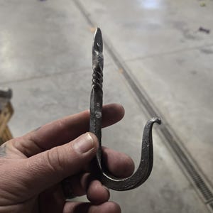 Puede incluir: Gancho de metal forjado a mano con una sección superior retorcida y un gancho inferior curvado. El gancho está hecho de metal oscuro y texturizado, probablemente hierro o acero, y está diseñado para colgar objetos.