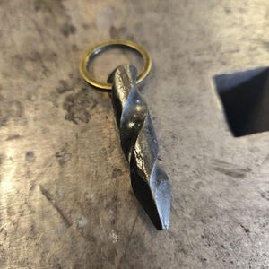 May include: A hand-forged metal keychain with a twisted design and a brass ring. The dark gray metal has a textured surface, and the keychain is designed for durability and style.
