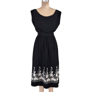 May include: A black vintage dress with a scoop neckline and cap sleeves. The dress features a fitted waist and a knee-length skirt. The hemline is adorned with a white floral embroidery design. The dress is displayed on a mannequin.