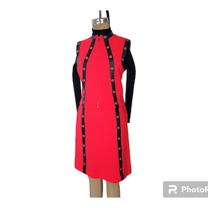 May include: A vintage red dress with black accents and gold-colored buttons. The dress features a high neckline and a zipper down the front. Black trim with gold buttons runs down the front and sides of the dress. The dress is displayed on a mannequin.