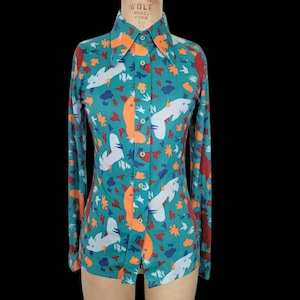 Peut inclure: Chemise à manches longues de couleur turquoise avec un motif répété de formes abstraites blanches, orange et rouges. La chemise a un col pointu et est boutonnée sur le devant.