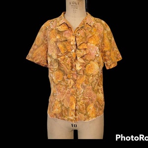 Puede incluir: Una blusa de manga corta con botones y cuello fruncido. La blusa está hecha de una tela ligera con estampado floral en tonos naranja, marrón y blanco.