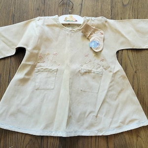 Op de afbeelding: Een vintage witte babyjurk met geborduurde bloemen en zakken. De jurk heeft een blauwe rand rond de halslijn en zoom. Een klein blauw label met de tekst "CHILDREN'S WEAR" is aan de jurk bevestigd.