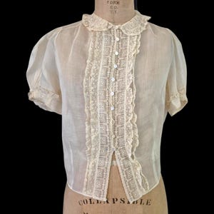 Puede incluir: Blusa vintage color crema con mangas cortas abullonadas y delicado ribete de encaje. La blusa presenta un cuello Peter Pan y un panel vertical de encaje en la parte delantera, asegurado con pequeños botones redondos. La tela parece transparente y ligera.