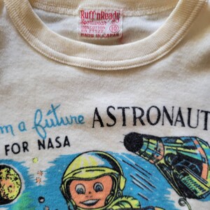 May include: Cream-colored t-shirt with a crew neck and a colorful graphic. The graphic features a cartoon astronaut, planets, and a spaceship, with the text "I'm a future ASTRONAUT FOR NASA."