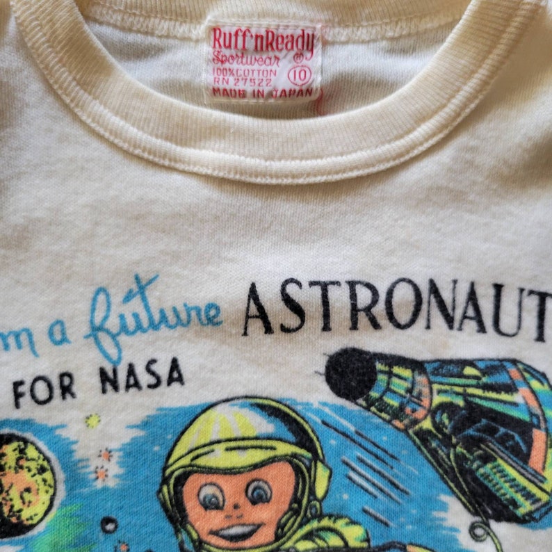 May include: Cream-colored t-shirt with a crew neck and a colorful graphic. The graphic features a cartoon astronaut, planets, and a spaceship, with the text "I'm a future ASTRONAUT FOR NASA."