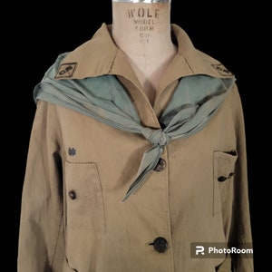 May include: A tan-colored uniform jacket with a green scarf tied around the neck. The jacket has two pockets and a button closure. The scarf has a dark blue emblem on it.