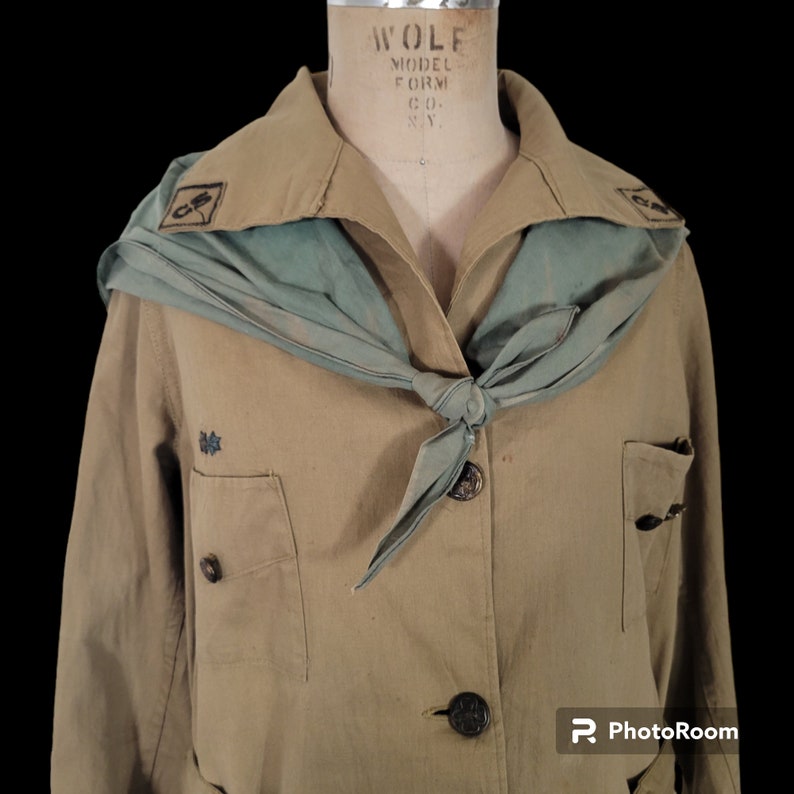 May include: A tan-colored uniform jacket with a green scarf tied around the neck. The jacket has two pockets and a button closure. The scarf has a dark blue emblem on it.