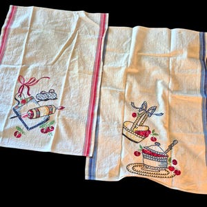 May include: Two off-white kitchen towels with embroidered designs. One towel features a cutting board, rolling pin, and cherries. The other has a basket, a pot with a lid, and cherries. Both have red and blue stripes.