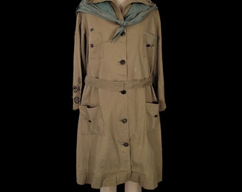 May include: A vintage Girl Scout uniform dress in a khaki color with a green sash and embroidered patches. The dress has a long sleeve, a button-up front, and a belt.