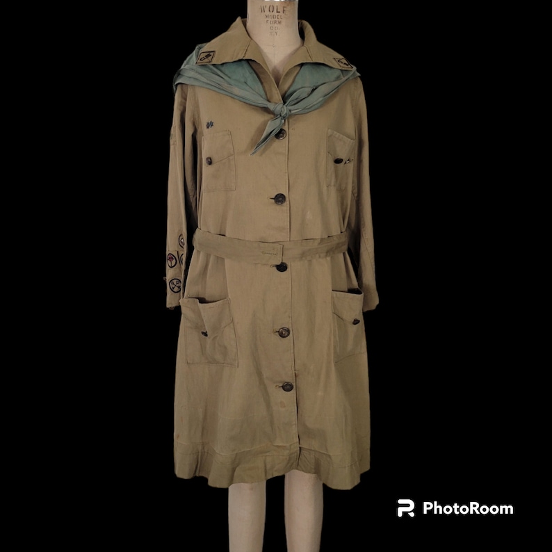 May include: A vintage Girl Scout uniform dress in a khaki color with a green sash and embroidered patches. The dress has a long sleeve, a button-up front, and a belt.
