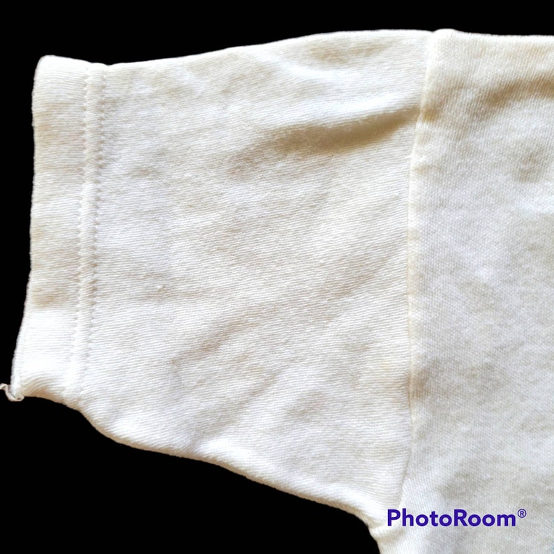 May include: Close-up of a cream-colored t-shirt sleeve. The sleeve's edge is neatly hemmed with a double-stitched seam. The fabric has a slightly textured appearance, and the overall look is clean and simple.