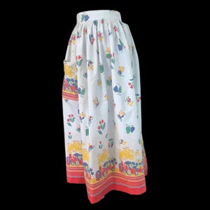 May include: A white gathered skirt featuring a repeating print of red steam locomotives with yellow smoke, interspersed with small red tulips and blue floral accents. The hem is finished with horizontal red and yellow stripes along the bottom edge.