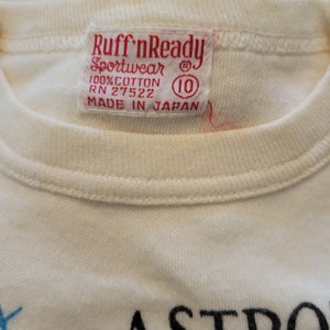 May include: Close-up of a cream-colored t-shirt with a red tag. The tag reads "Ruff'nReady Sportswear" and "100% Cotton" with the number "10" in a circle. The shirt is made in Japan.