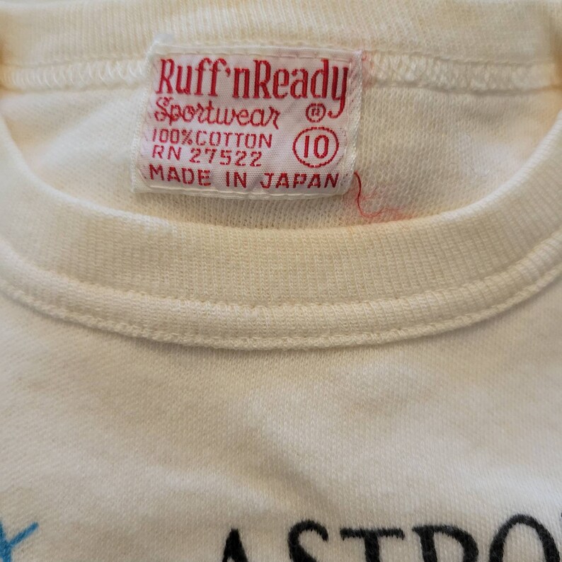 May include: Close-up of a cream-colored t-shirt with a red tag. The tag reads "Ruff'nReady Sportswear" and "100% Cotton" with the number "10" in a circle. The shirt is made in Japan.
