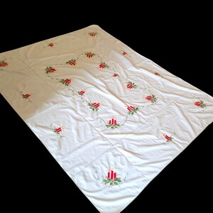 Op de afbeelding: Witte tafelkleed met geborduurde rode kaarsen en groene hulstbladeren. Het tafelkleed is gemaakt van een zachte, lichte stof en is perfect voor een feestelijk diner.