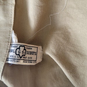 May include: A tan fabric label with black text that reads "TRADE MARK GIRL GS SCOUTS REG. U. S. PAT. OFF. GIRL SCOUTS INC. NEW YORK CITY".