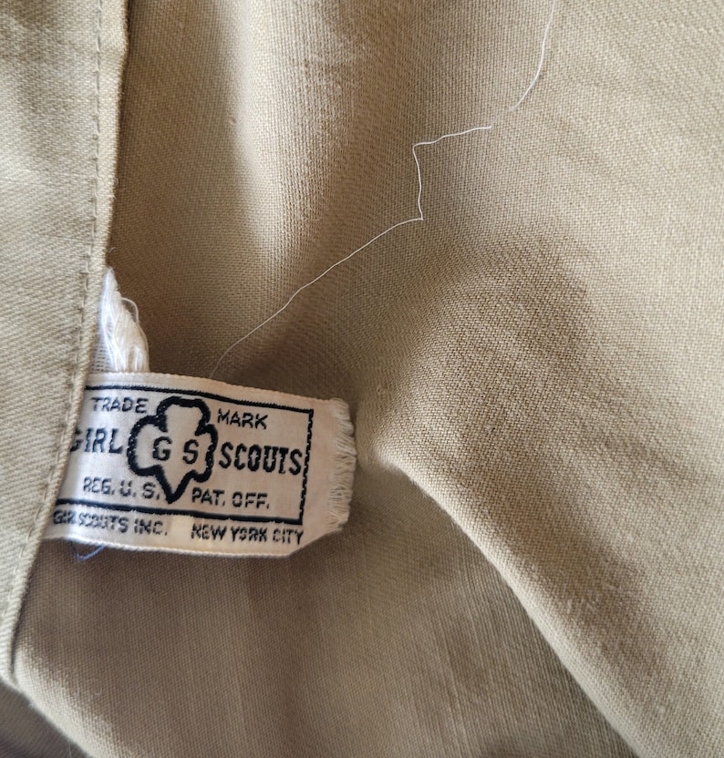 May include: A tan fabric label with black text that reads "TRADE MARK GIRL GS SCOUTS REG. U. S. PAT. OFF. GIRL SCOUTS INC. NEW YORK CITY".