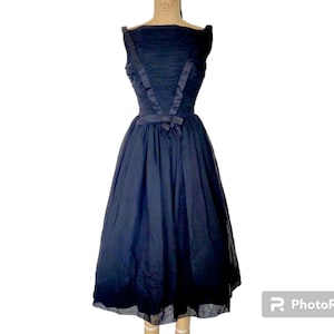 Può includere: Un abito in tulle blu navy senza maniche con un corpetto aderente e una gonna ampia. Il corpetto presenta un dettaglio drappeggiato e un fiocco di raso nero in vita.