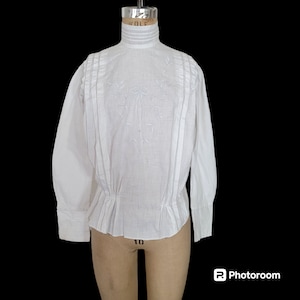 May include: White cotton blouse with a high neck and long sleeves. The blouse has a gathered waist and is decorated with white embroidery.