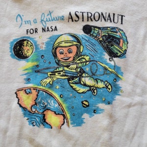 May include: Off-white t-shirt with a retro illustration of an astronaut in a yellow spacesuit, floating in space. The design includes the text "I'm a future ASTRONAUT FOR NASA", a planet, and a spaceship.