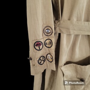 May include: A beige utility jacket with a variety of embroidered patches on the sleeve. The patches feature symbols like an eye, a tree, a bed, a key, and an owl.