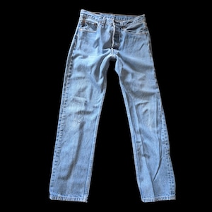May include: Light blue denim jeans featuring a classic five-pocket design, a button fly closure, and a straight-leg cut. The fabric displays a faded wash with subtle whiskering details at the hips and a small distressed mark on the left thigh.